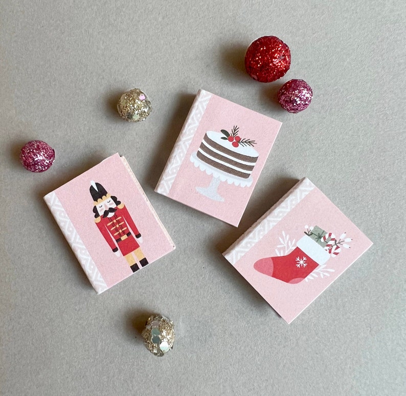May include: Four pink gift boxes with different holiday designs. The boxes feature a nutcracker, a cake, a stocking, and a candy cane.