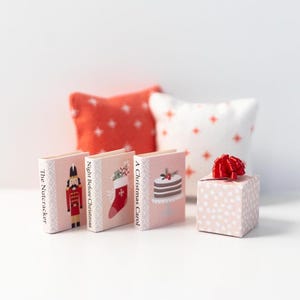 May include: Three miniature books with Christmas themes: "The Nutcracker", "Night Before Christmas", and "A Christmas Carol". Each book has a different illustration on the cover. A small pink gift box with a red bow is in the foreground.
