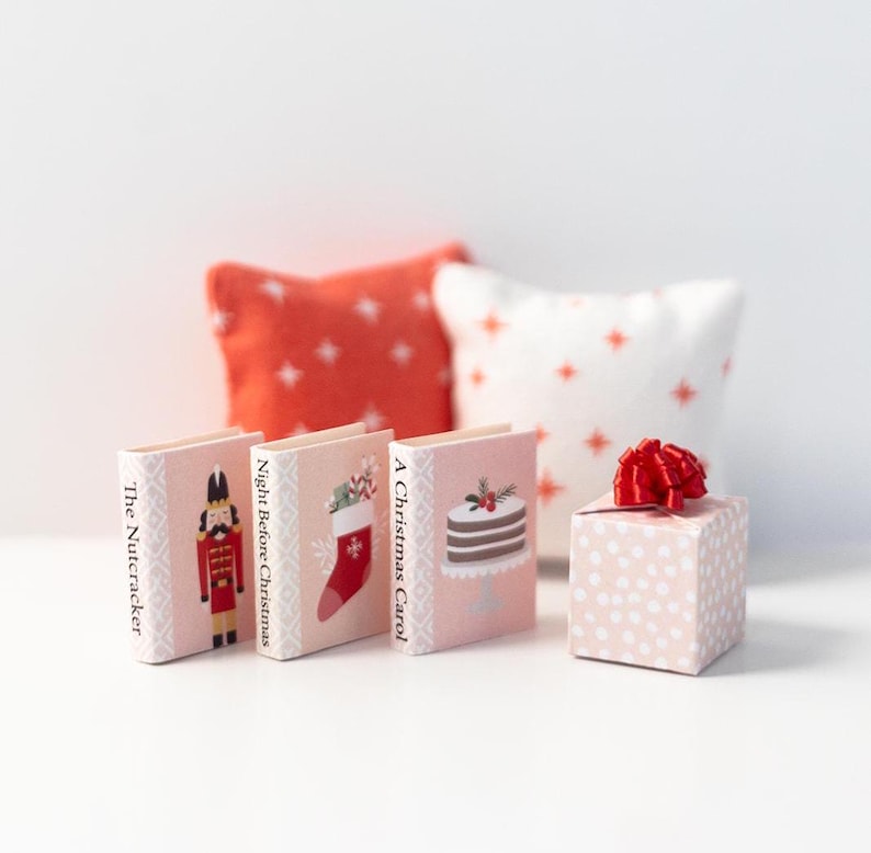 May include: Three miniature books with Christmas themes: "The Nutcracker", "Night Before Christmas", and "A Christmas Carol". Each book has a different illustration on the cover. A small pink gift box with a red bow is in the foreground.