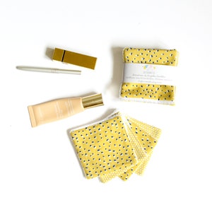 May include: An overhead shot features beauty products and reusable facial rounds. A gold lipstick case and a silver eyeliner pencil are next to a tube of foundation. A stack of yellow and navy spotted facial rounds is visible.