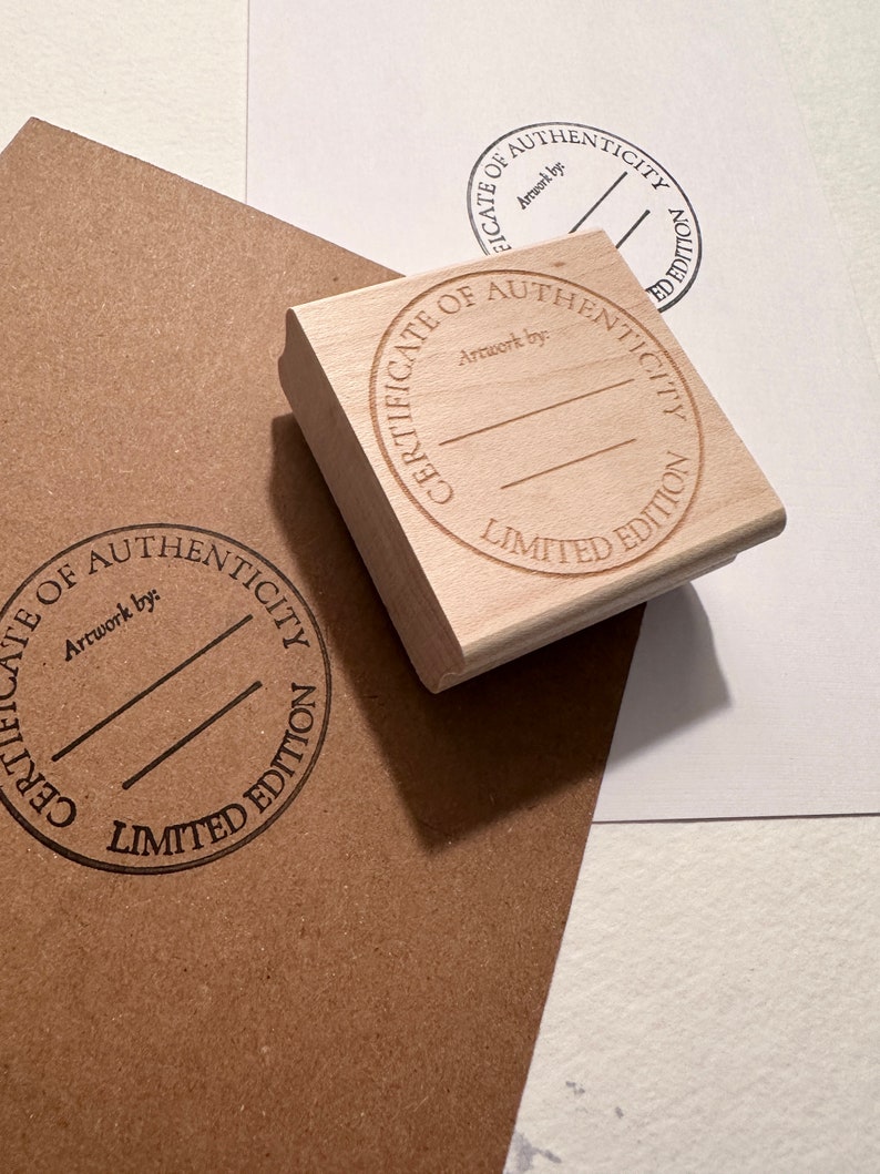 May include: A wooden stamp with the text "Certificate of Authenticity Limited Edition" and the words "Artwork by:" in a circle. The stamp is on a brown paper surface.