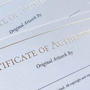 May include: Close-up of two white certificates of authenticity. The text "CERTIFICATE OF AUTHENTICITY" is printed in gold foil. The text "Original Artwork By:" is printed in a dark blue font. The bottom of the certificate has a signature.