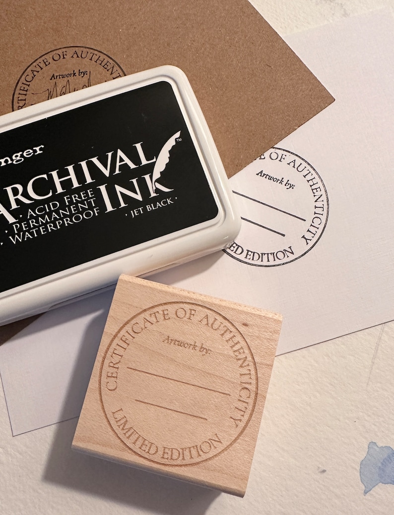 May include: Two wooden stamps with the text "Certificate of Authenticity Limited Edition" and "Certificate of Authenticity" with a blank space for a signature. The stamps are on a brown paper surface.