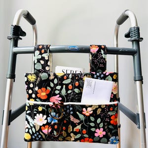 May include: A black fabric walker bag featuring a colorful floral and strawberry print with two top straps for attachment. The bag includes two front pockets containing a white envelope and a pair of tortoiseshell glasses, with a white trim.