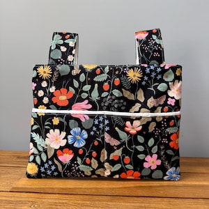 May include: A rectangular fabric tote bag featuring a black background with a colorful botanical print of red strawberries, various wildflowers, and small moths. The bag includes two top handles and a horizontal white trim across the front pocket.