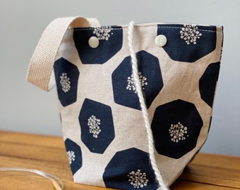 Pode incluir: Uma bolsa tote de tecido bege e preto com fecho de cordão e uma alça de algodão branco. A bolsa tem um padrão geométrico de hexágonos pretos com desenhos florais brancos.