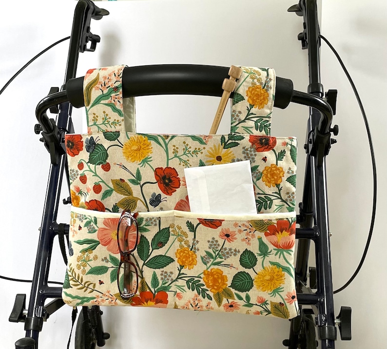 Puede incluir: Bolsa para andador con estampado floral sobre fondo crema, con amapolas rojas, margaritas amarillas y hojas verdes. La bolsa tiene dos bolsillos, un par de gafas y un paquete de pa&ntilde;uelos. Se ven agujas de tejer de madera. La bolsa est&aacute; sujeta a un andador negro.