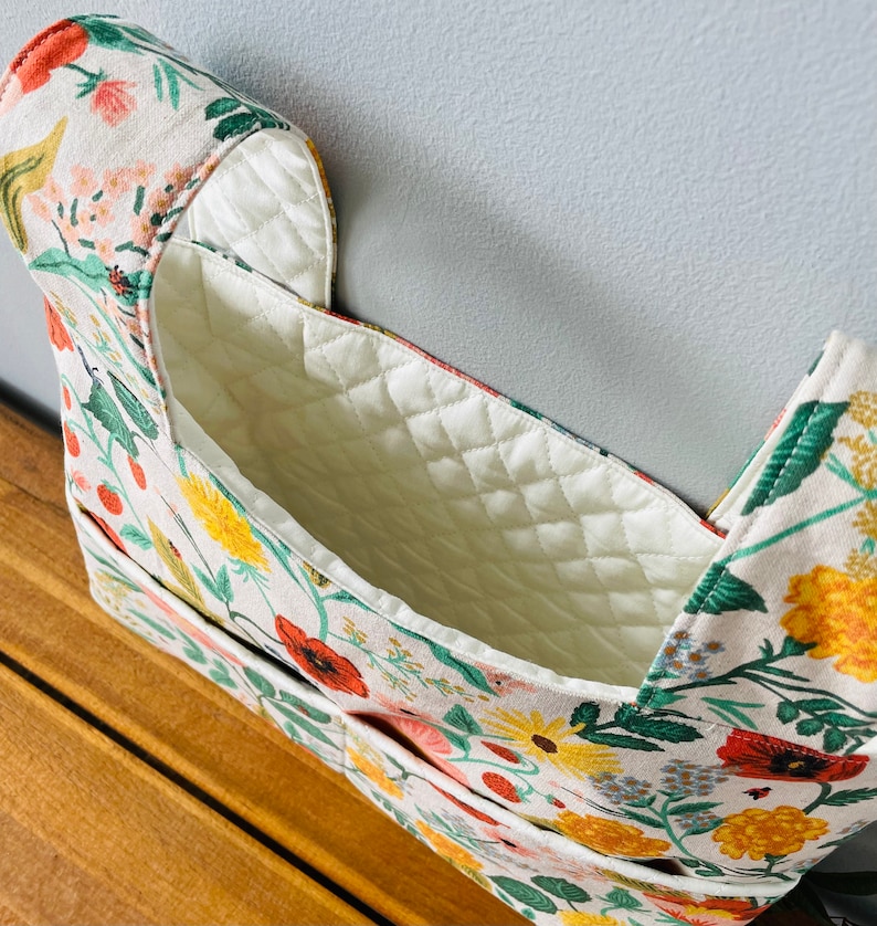 Puede incluir: Una bolsa organizadora con estampado floral con un interior acolchado de color crema. El exterior presenta una impresi&oacute;n vibrante de amapolas rojas, margaritas amarillas y follaje verde sobre un fondo crema. La bolsa tiene m&uacute;ltiples bolsillos y un dise&ntilde;o estructurado.