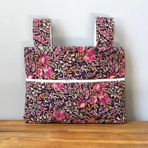 May include: A rectangular fabric bag featuring a dark purple background with a dense floral pattern in shades of pink, orange, and white. The bag includes two top handles and a horizontal front pocket accented with a white trim along the top edge.