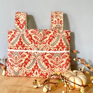 May include: A rectangular fabric bag featuring a burnt orange and cream damask pattern. The bag has two upright fabric handles and a horizontal front pocket with a white trim edge. The textile displays a repeating ornate floral motif.