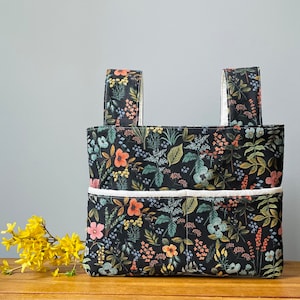 May include: A rectangular fabric tote bag featuring a black background with a colorful botanical print of flowers and leaves in shades of pink, yellow, blue, and green. The bag has two top handles and a front pocket with a white piped trim.
