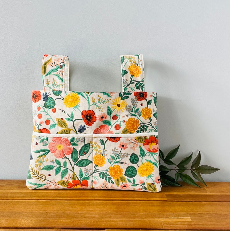 Puede incluir: Un bolso tote rectangular con dos asas, con un estampado floral con flores rojas, amarillas y rosas, hojas verdes y mariposas sobre un fondo crema. El bolso tiene dos bolsillos delanteros y est&aacute; sobre una superficie de madera.