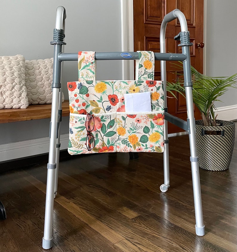 Puede incluir: Bolsa para andador con estampado floral con m&uacute;ltiples bolsillos, sujeta a un andador plateado. La bolsa presenta un fondo crema con estampados florales rojos, amarillos y verdes. Un par de gafas y papeles blancos est&aacute;n en los bolsillos.