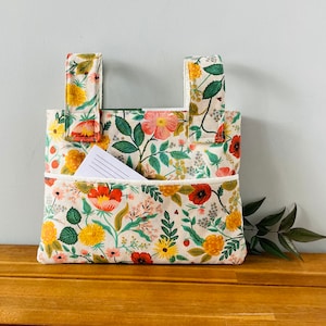 Puede incluir: Bolso tote con estampado floral sobre fondo crema, con flores rojas, amarillas y rosas y hojas verdes. El bolso tiene dos asas y un bolsillo delantero con un bloc de notas rayado en su interior. El bolso est&aacute; sobre una superficie de madera.