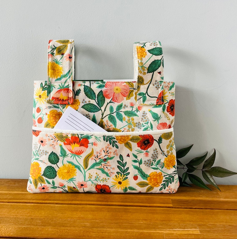 Puede incluir: Bolso tote con estampado floral sobre fondo crema, con flores rojas, amarillas y rosas y hojas verdes. El bolso tiene dos asas y un bolsillo delantero con un bloc de notas rayado en su interior. El bolso est&aacute; sobre una superficie de madera.