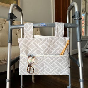 May include: A gray and white geometric patterned fabric storage pouch attached to a silver metal walker. The pouch features two top straps and a front pocket containing a pair of tortoiseshell-rimmed eyeglasses and two wooden knitting needles.