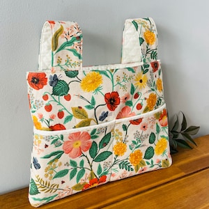 Puede incluir: Un bolso tote con estampado floral con dos bolsillos y dos asas. El bolso presenta un fondo crema con amapolas rojas, flores amarillas, hojas verdes y fresas. Las asas y el ribete de los bolsillos son blancos.