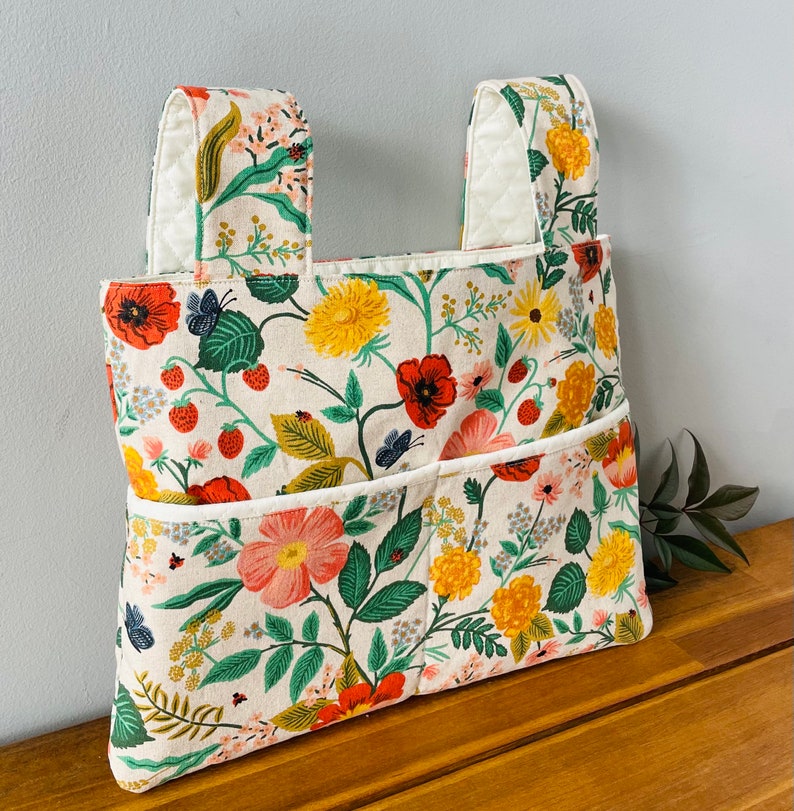 Puede incluir: Un bolso tote con estampado floral con dos bolsillos y dos asas. El bolso presenta un fondo crema con amapolas rojas, flores amarillas, hojas verdes y fresas. Las asas y el ribete de los bolsillos son blancos.
