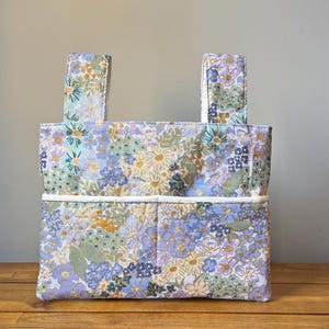 May include: A rectangular fabric tote bag featuring a repeating floral pattern in shades of light blue, lavender, yellow, and green. The bag includes two upright fabric handles and two exterior pockets across the front with white trim.