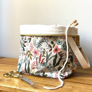 May include: A floral print craft bag with a cream-colored interior and a beige strap. The bag features a zipper closure and a gold-colored zipper. Knitting needles and scissors are visible. The bag is designed for carrying craft supplies.