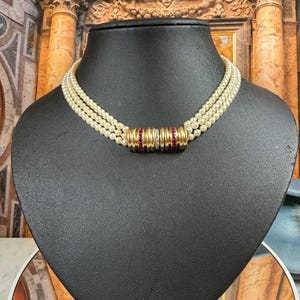 Op de afbeelding: Een parelketting met drie strengen, gouden accenten en rode edelstenen. De ketting wordt getoond op een zwarte mannequin. De gouden accenten zijn cilindrisch en omcirkelen de ketting in het midden. De achtergrond toont architecturale details.
