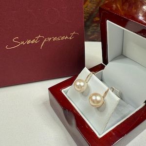 May include: Gold-coloured drop earrings with large, round, peach-coloured pearls, presented in an open red wooden jewellery box. The earrings rest on a white cushion. A red gift box with the words "Sweet present" is visible.