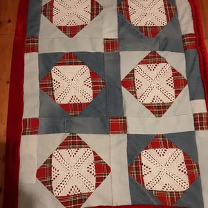 Può includere: Trapunta fatta a mano con un design patchwork. Incorpora quadrati di denim, accenti scozzesi rossi e verdi e centrotavola all'uncinetto bianchi. Un bordo rosso in peluche incornicia la trapunta, creando un tessuto decorativo.