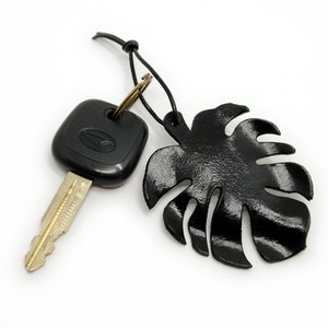 May include: A black leather monstera leaf keychain with a black car key attached. The keychain has a black cord and a gold metal ring.