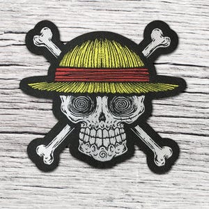 Könnte beinhalten: Ein gestickter Aufnäher mit einem weißen Totenkopf mit Spiralaugen, der einen gelben Strohhut mit rotem Band trägt, platziert über zwei gekreuzten weißen Knochen auf schwarzem Hintergrund. Das Design ist ein klassisches Piraten-Emblem mit Stickerei.