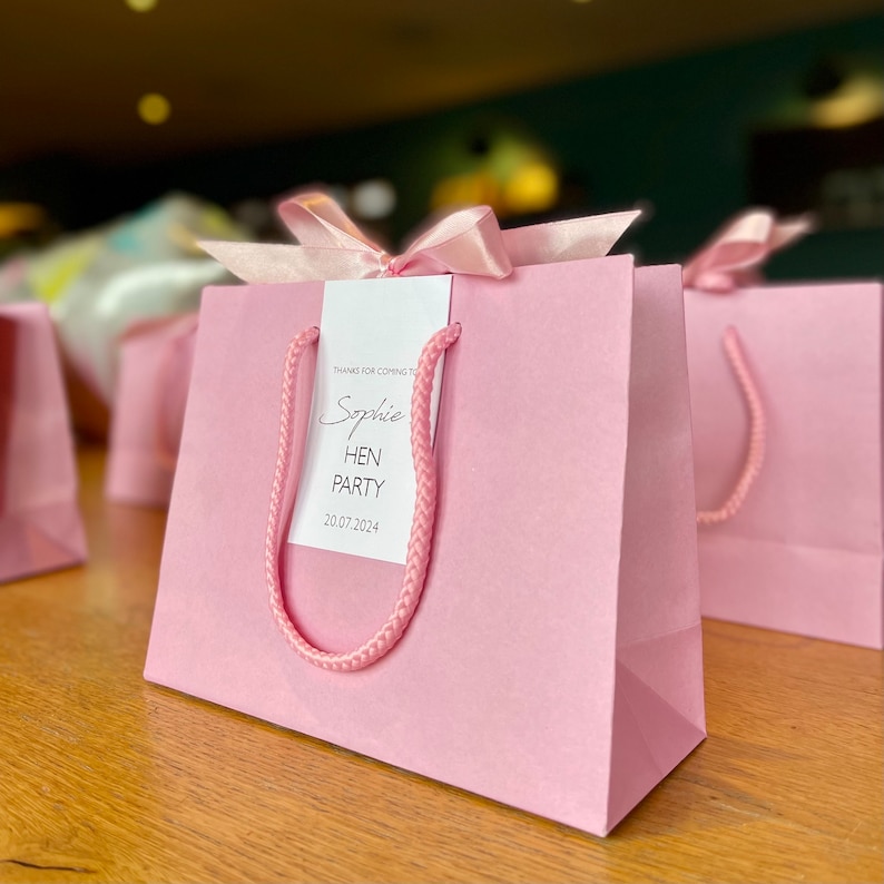 May include: Pink gift bags with matching rope handles and a satin ribbon bow. Each bag has a tag that reads "Sophie HEN PARTY 20.07.2024". The bags are on a wooden surface.