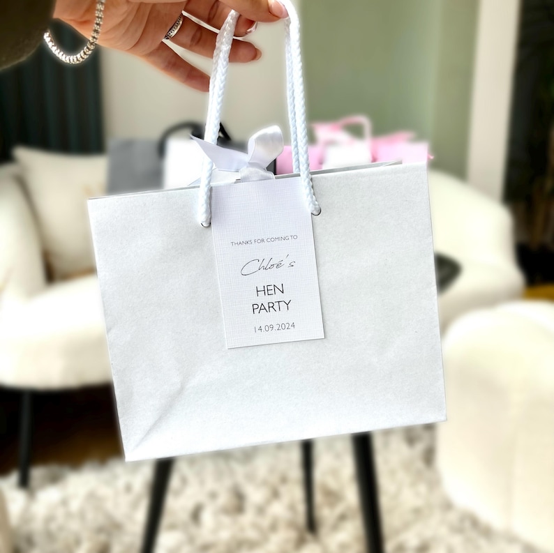 May include: A white gift bag with white rope handles and a white tag. The tag reads "Thanks for coming to Chloe's Hen Party 14.09.2024". A white ribbon is tied to the top of the bag. The bag is held by a person.