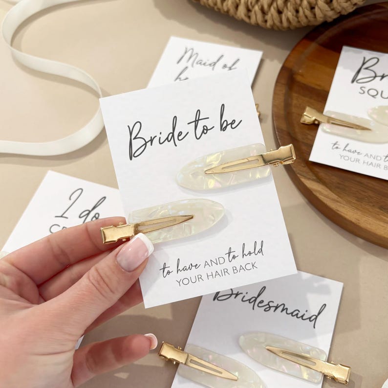 May include: Set of four hair clips with white pearlescent accents and gold metal clasps. Each clip has a card attached with text that reads "Bride to be", "Maid of Honour", "Bridesmaid", and "Bride Squad".