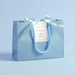 May include: A light blue gift bag with a matching ribbon and tag. The tag reads "Paige's HEN PARTY 07.11.2025" in elegant script. The bag has a twisted cord handle, perfect for party favors.