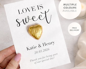 May include: A white card with the text "LOVE IS sweet" and the names "Katie & Henry" with the date "20.03.2026". A gold foil-wrapped heart-shaped chocolate sits on the card. The card reads "Thank you for being part of our special day!"