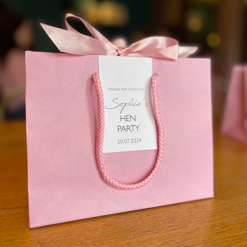 Personalised hen party gift bags with custom tags and ribbons, perfect for adding a special touch to hen do favours. Made from 100% recycled paper with reinforced bases and soft rope handles, these elegant bags can be customised with names, dates, or messages. Ideal for presenting gifts or party favours in style.