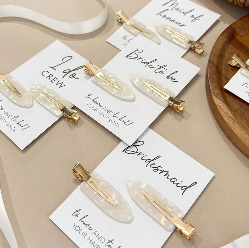May include: White and gold hair clips with iridescent accents are displayed on cards with wedding-related text. The cards read "I do crew", "Bride to be", "Bridesmaid", and "Maid of honour". A white ribbon is visible in the background.