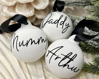 May include: Three round white ceramic ornaments with black satin ribbons for hanging. Each ornament features black cursive text reading Mummy, Daddy, and Arthur, respectively, displayed on the smooth, matte white surface of the spherical baubles.