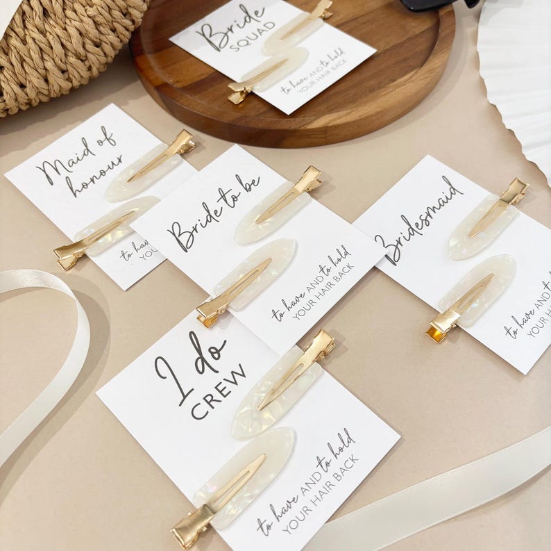 May include: A collection of bridal party hair clips with gold accents and white pearlescent oval designs. Each clip is paired with a card that has a title such as "Bride Squad" or "Maid of Honor". A white ribbon and a wooden tray are also in the image.