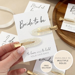 May include: Two pearlescent white hair clips with gold-colored metal clasps are displayed on a white card that reads "Bride to be". The card also has the text "to have and to hold YOUR HAIR BACK". The image suggests a bridal theme.
