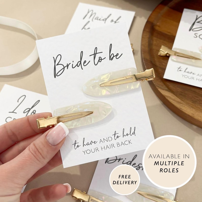 May include: Two pearlescent white hair clips with gold-colored metal clasps are displayed on a white card that reads "Bride to be". The card also has the text "to have and to hold YOUR HAIR BACK". The image suggests a bridal theme.