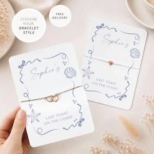 May include: Two white rectangular display cards featuring blue line art of seashells and starfish. One card holds a gold-tone chain bracelet with two interlocking heart charms, while the other holds a white string bracelet with a single heart charm.