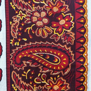 May include: Close-up of a textile with a paisley and floral pattern. The design features a deep burgundy background with red, yellow, and white accents. The pattern includes paisley shapes, flowers, and decorative borders.
