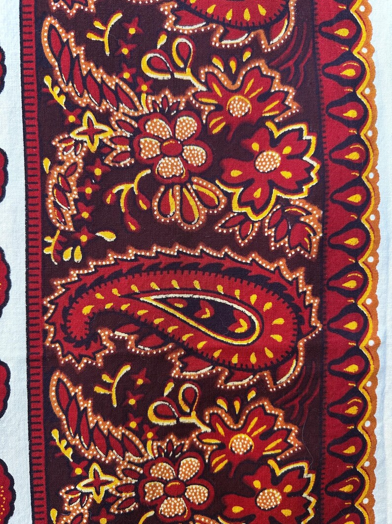 May include: Close-up of a textile with a paisley and floral pattern. The design features a deep burgundy background with red, yellow, and white accents. The pattern includes paisley shapes, flowers, and decorative borders.