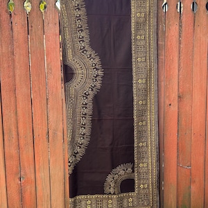 May include: A dark brown fabric with intricate gold patterns hangs on a wooden rod. The fabric features detailed, symmetrical designs along the edges and a large central motif. The textile is displayed against a weathered, orange wooden fence.