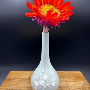 May include: A light blue ceramic vase with a white floral design, holding a single red and orange sunflower.