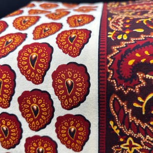 May include: Close-up of a fabric with a repeating pattern of red and yellow teardrop shapes on a white background. To the right is a section with a paisley design in red, yellow, and brown. The fabric is likely used for home decor.
