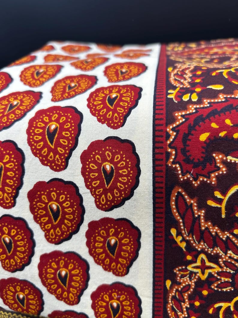 May include: Close-up of a fabric with a repeating pattern of red and yellow teardrop shapes on a white background. To the right is a section with a paisley design in red, yellow, and brown. The fabric is likely used for home decor.