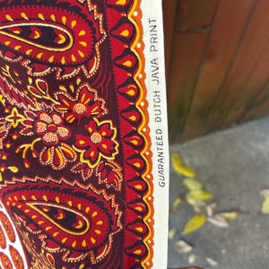 May include: Close-up of a fabric with a paisley design in shades of red, brown, and yellow. The fabric has a white border with the text "GUARANTEED DUTCH JAVA PRINT". The pattern includes floral and teardrop shapes.