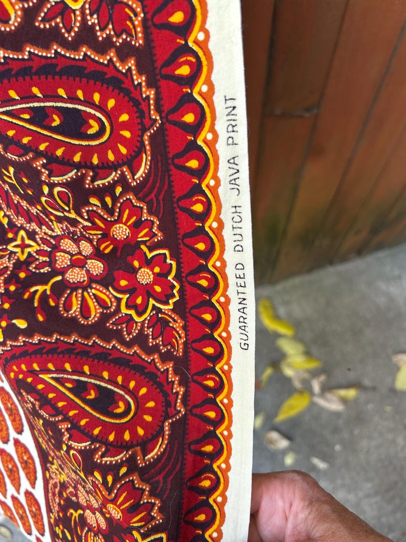 May include: Close-up of a fabric with a paisley design in shades of red, brown, and yellow. The fabric has a white border with the text "GUARANTEED DUTCH JAVA PRINT". The pattern includes floral and teardrop shapes.