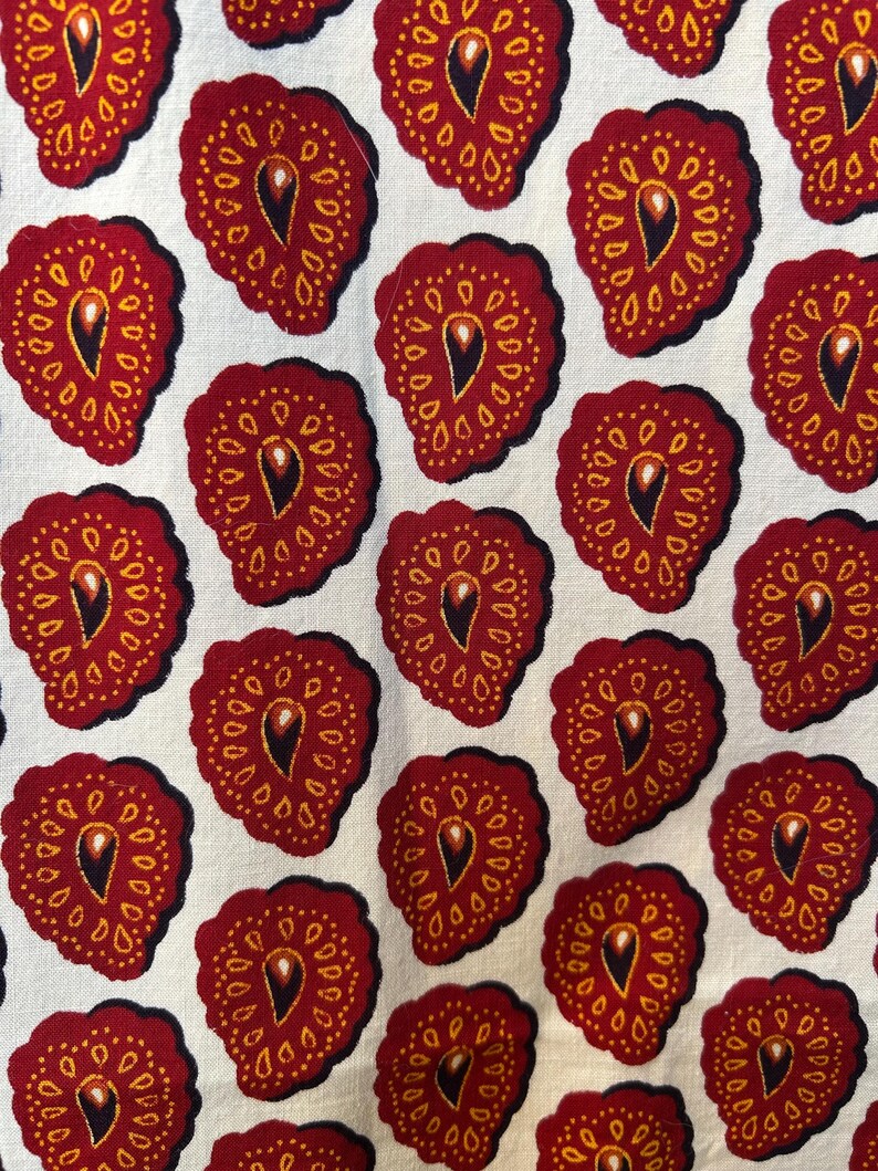 May include: Close-up of a fabric with a repeating pattern of stylized red fruit shapes. Each shape has a dark outline, a yellow dotted border, and a central teardrop design. The background is a light cream color.